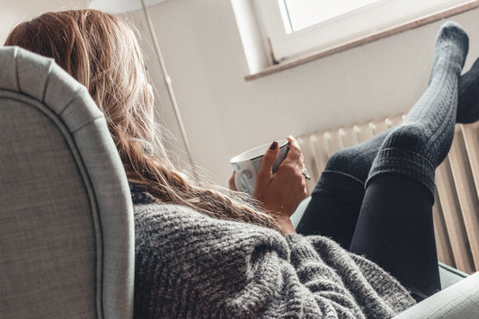 Woman wearing cosy thermal wear including over-knee socks with the feet on the radiator holding a mug of hot beverage 