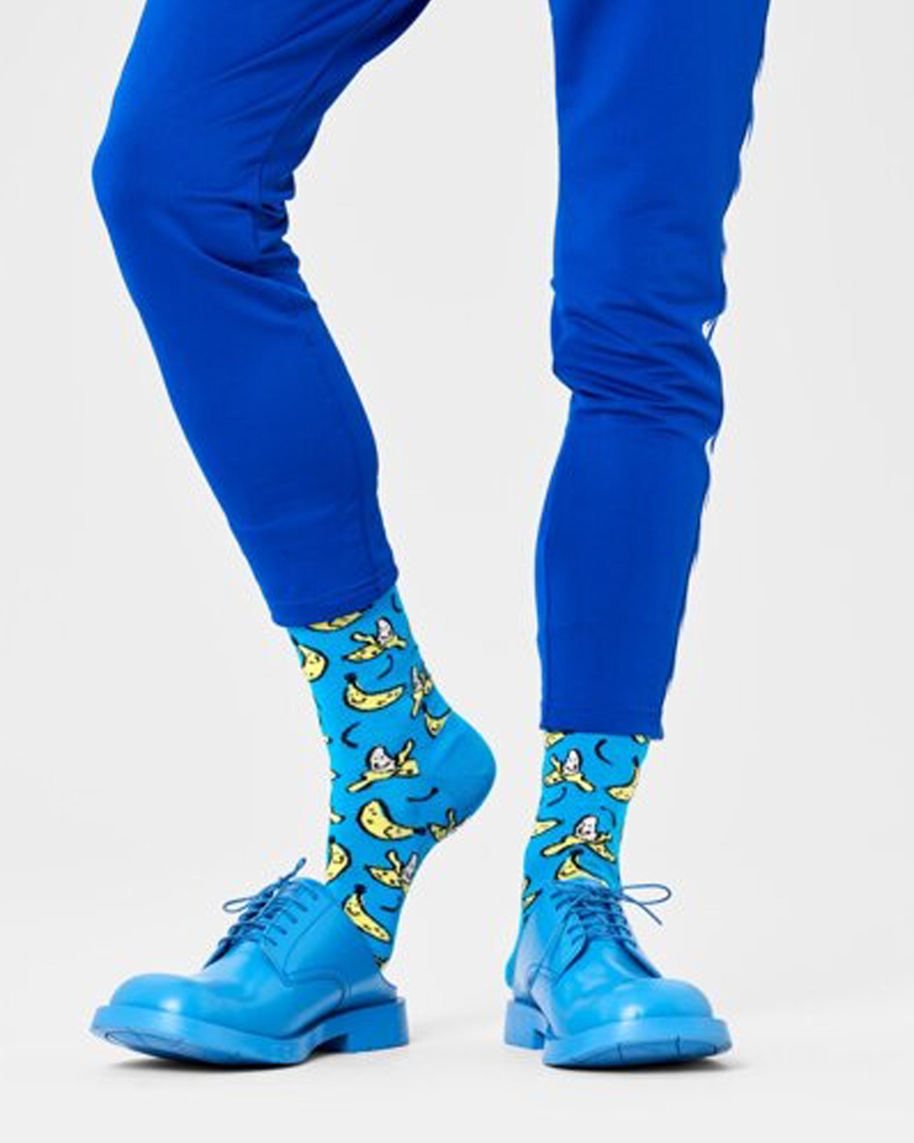 Happy Socks P000720 Banana Sock - Blue cotton crew length ankle socks with an all over pattern of smiling cartoon bananas in yellow, cream and black, worn with blue brogues and blue pants.