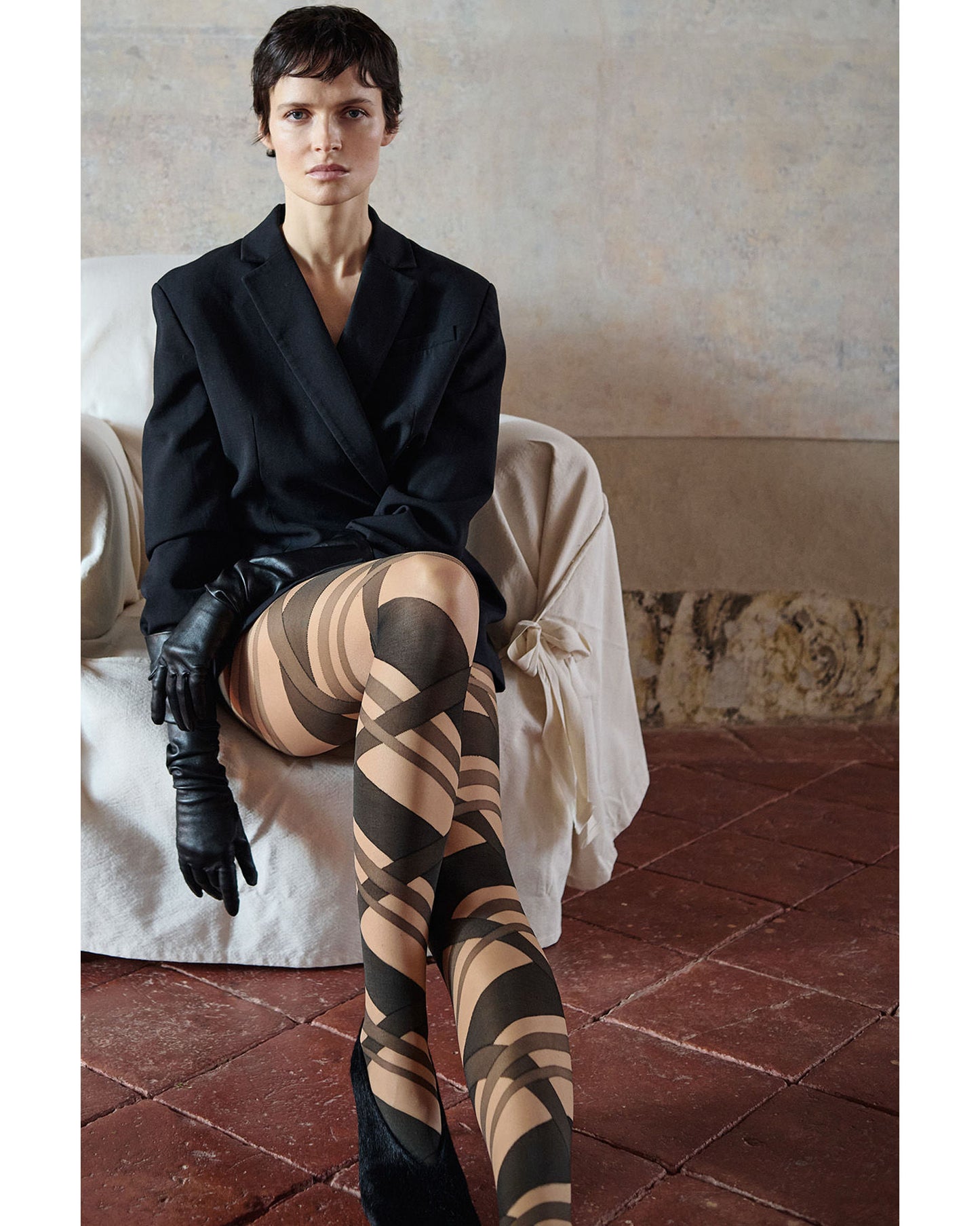 Woman wearing a black blazer, tan and black striped patterned tights, and black gloves sitting on a white chair.