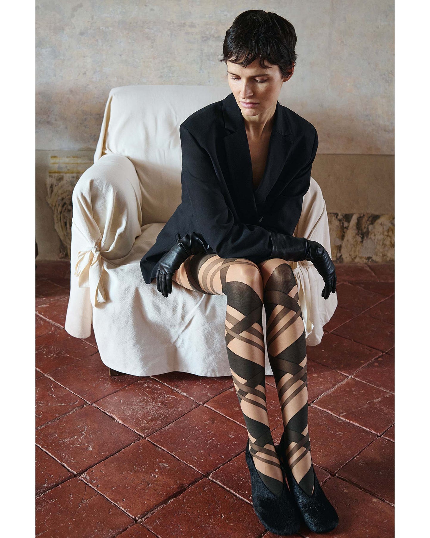 Woman sitting on a white chair wearing a black blazer, black gloves and nude and black striped patterned tights.