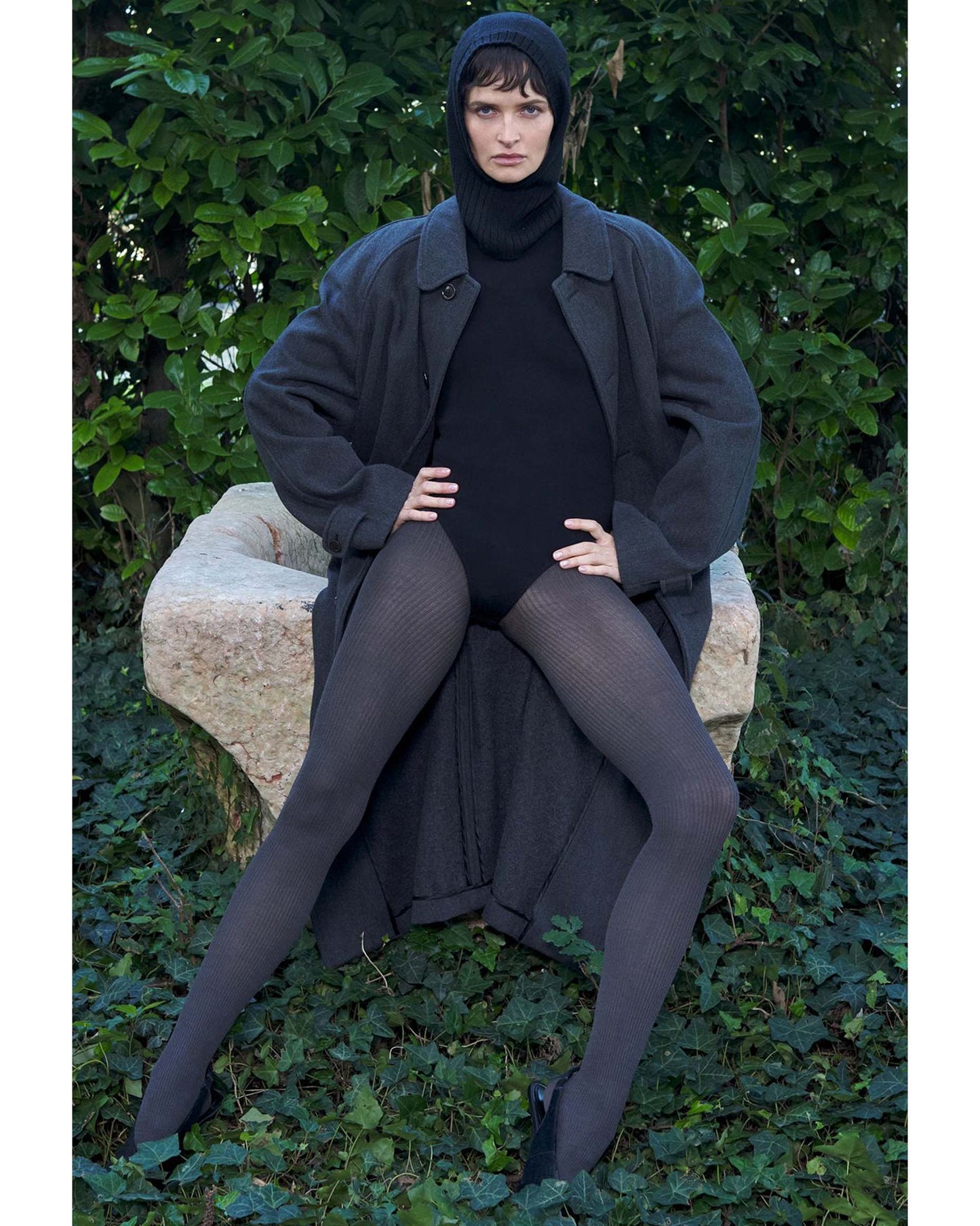 Person wearing a long dark coat, black turtleneck bodysuit and grey ribbed tights sitting on a stone bench with green foliage in the background