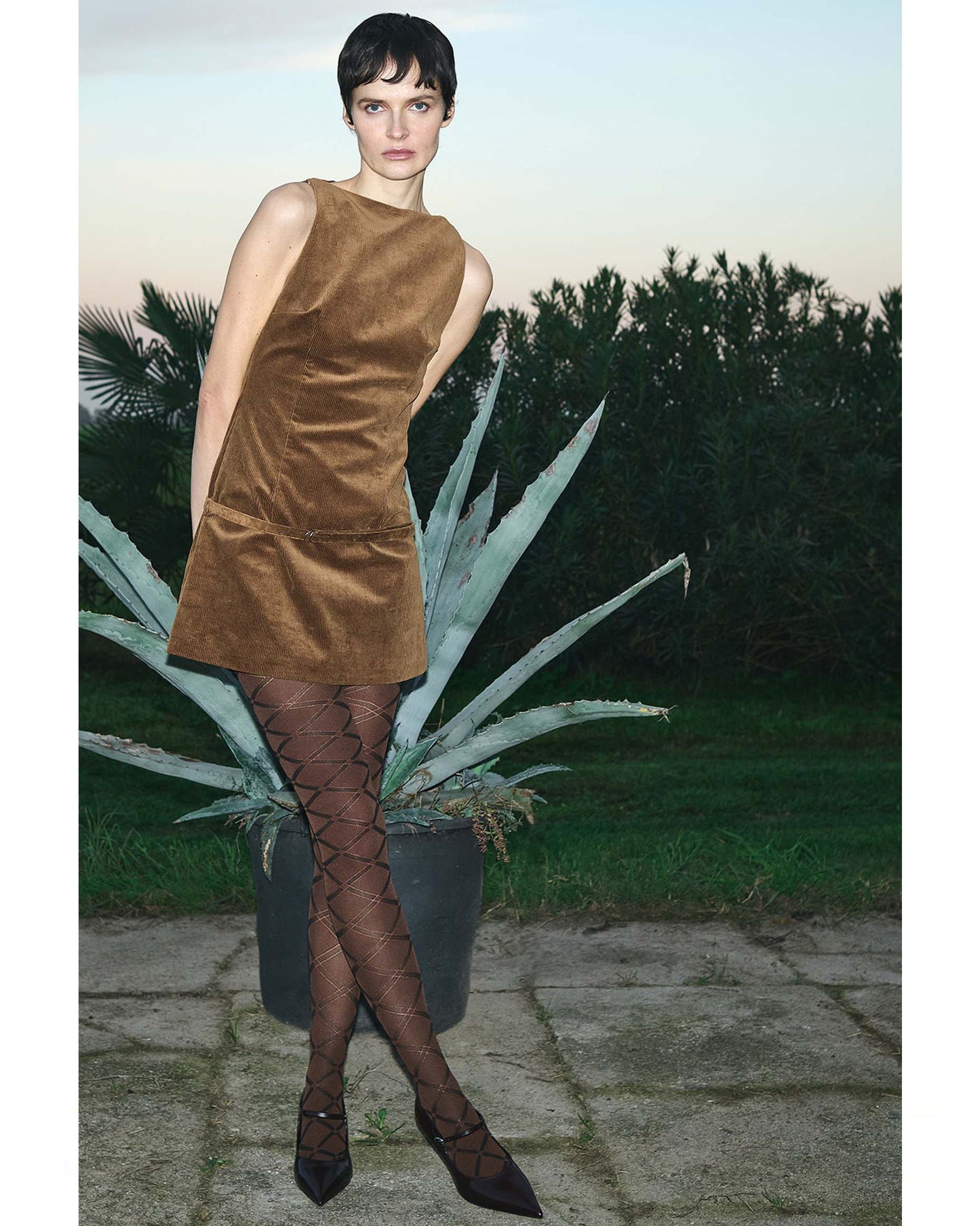 Woman in a brown dress and patterned stockings standing outdoors with plants in the background