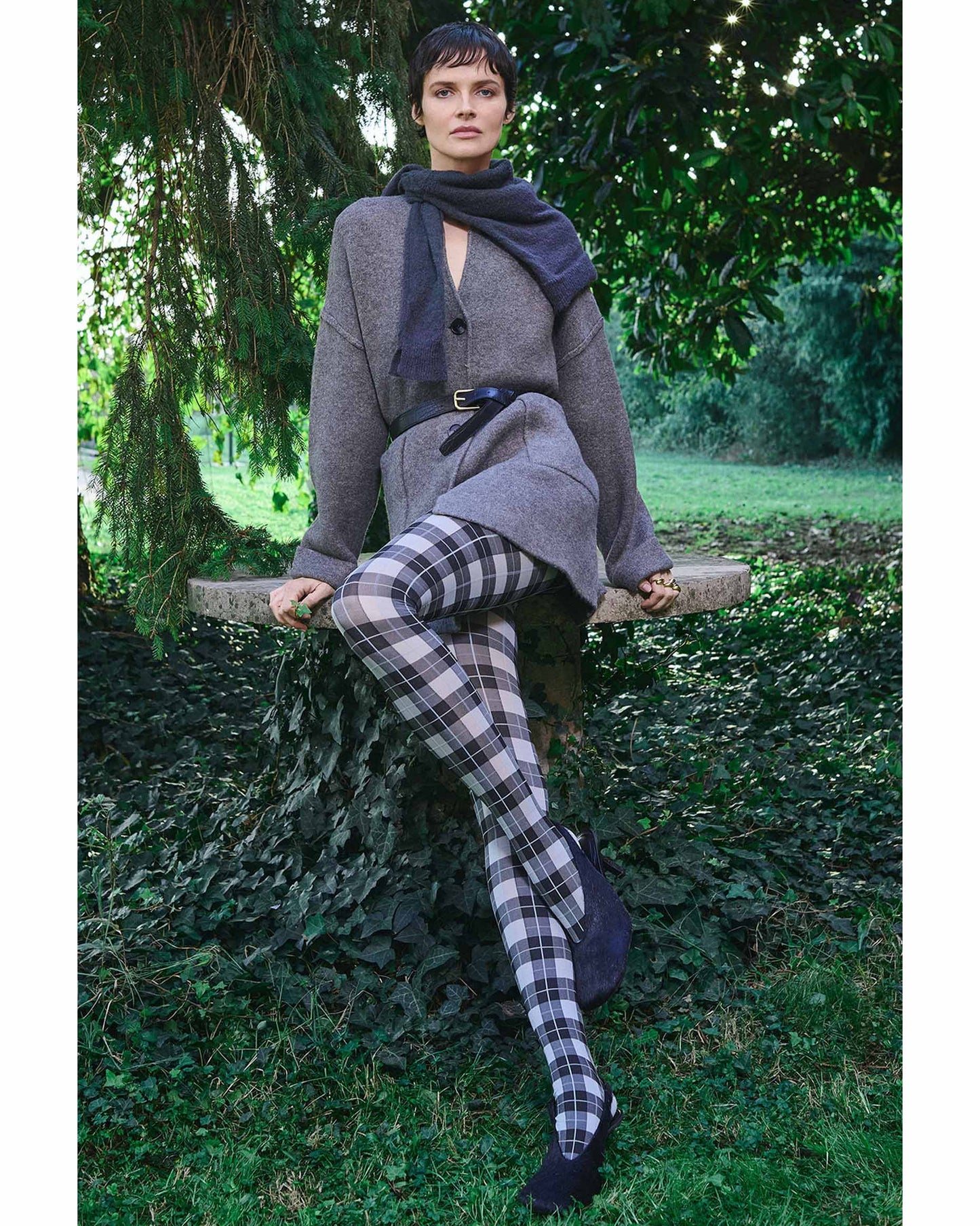 Woman sitting outdoors wearing a gray coat, plaid/tartan tights, and black shoes.