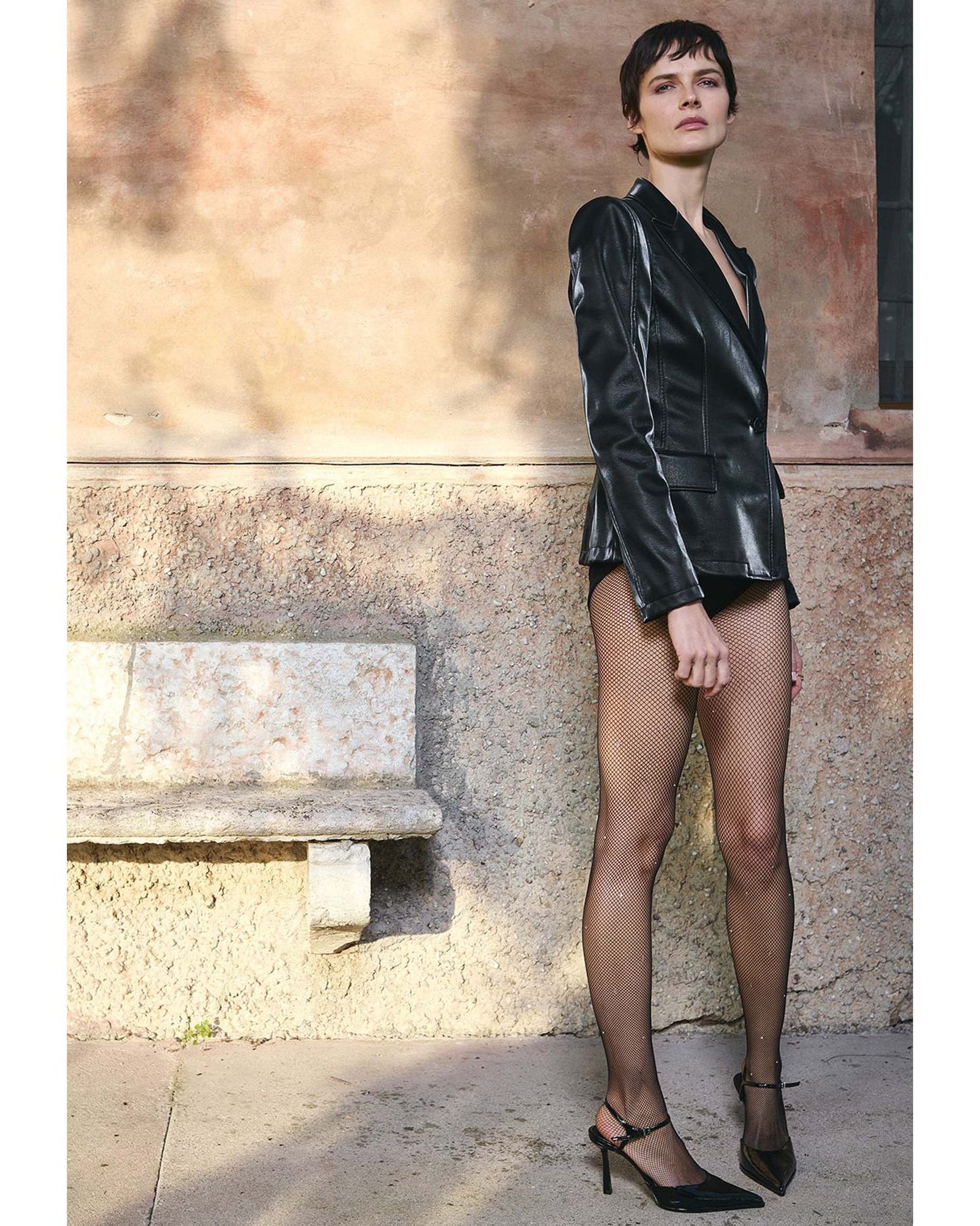 Woman in a black outfit and black crystal fishnets standing against a stone wall.