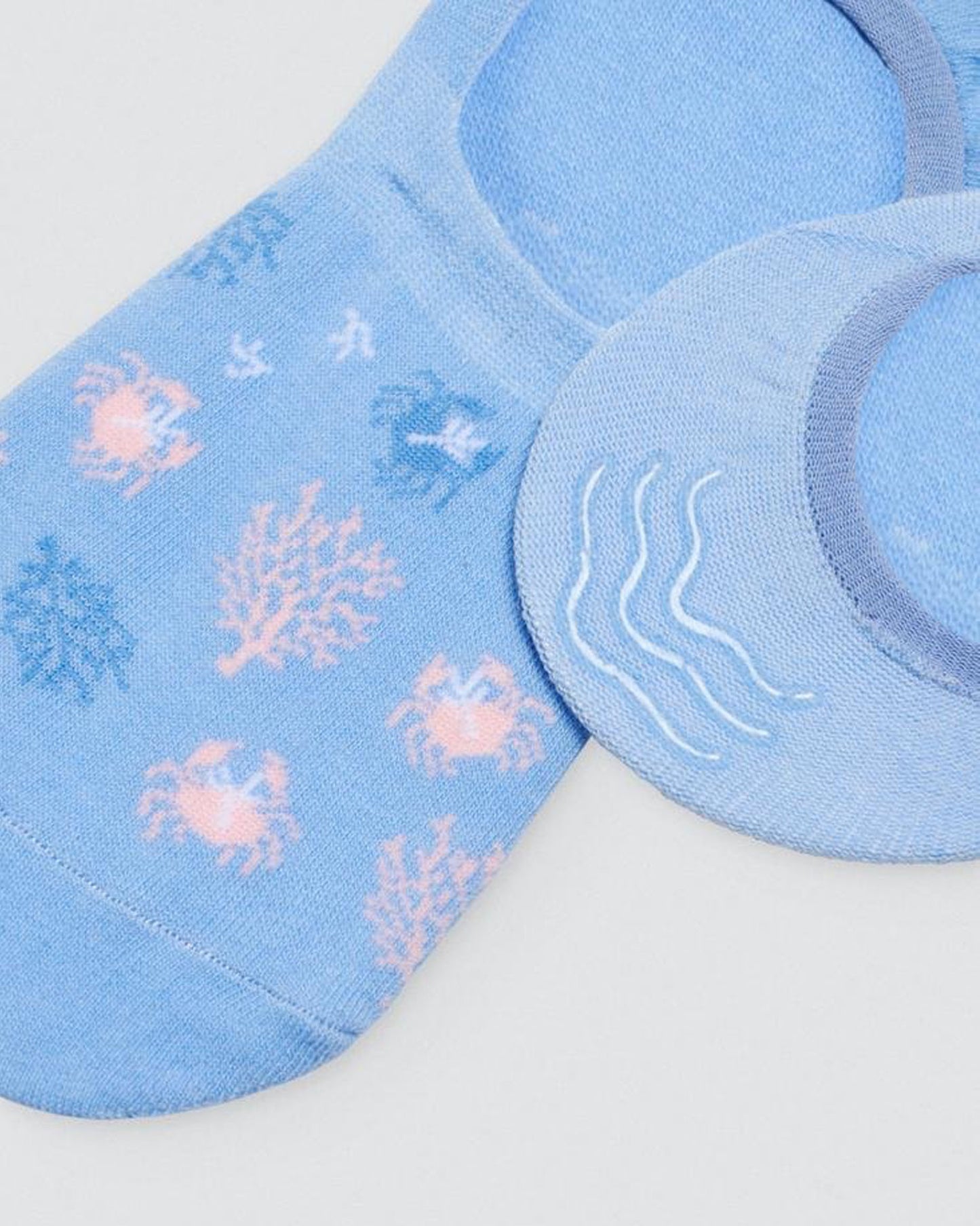 Ysabel Mora Crabs & Coral Shoe Liners - Light blue cotton no show shoe liner sock with crabs and coral pattern in pale pink/peach and blue and three strips of silicone on the heel. Close up detail.