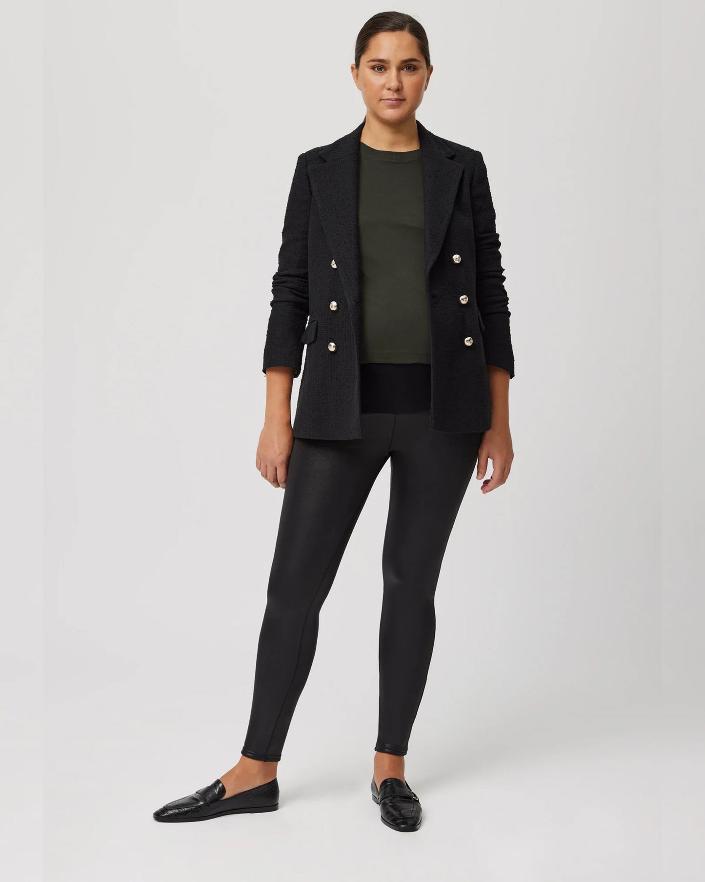 Pregnant woman wearing a black blazer with gold buttons, khaki green top, black waxed look pants and black shoes on a white background
