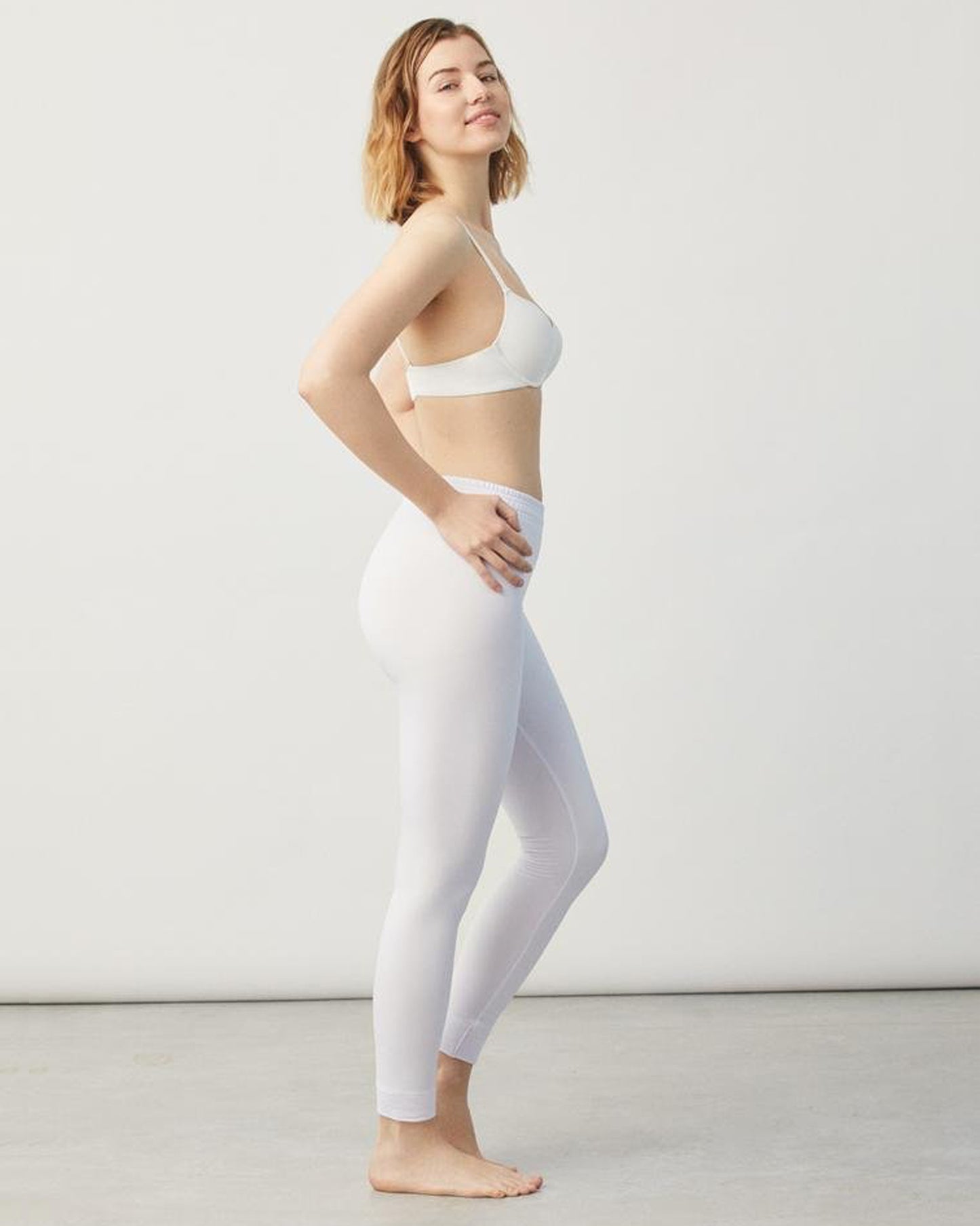 Woman wearing a white sports bra and leggings on a plain background, side view