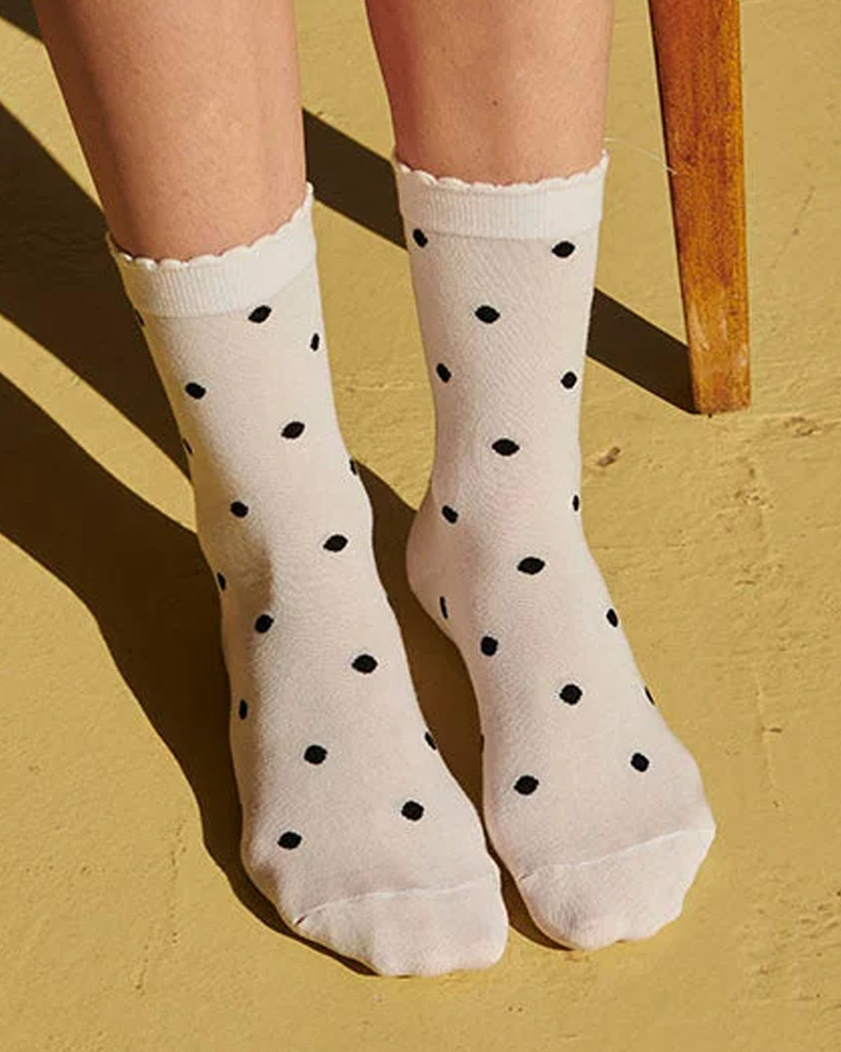 A pair of off-white crew socks with black polka dots worn on feet, shown against a yellow wall with a wooden chair leg visible.