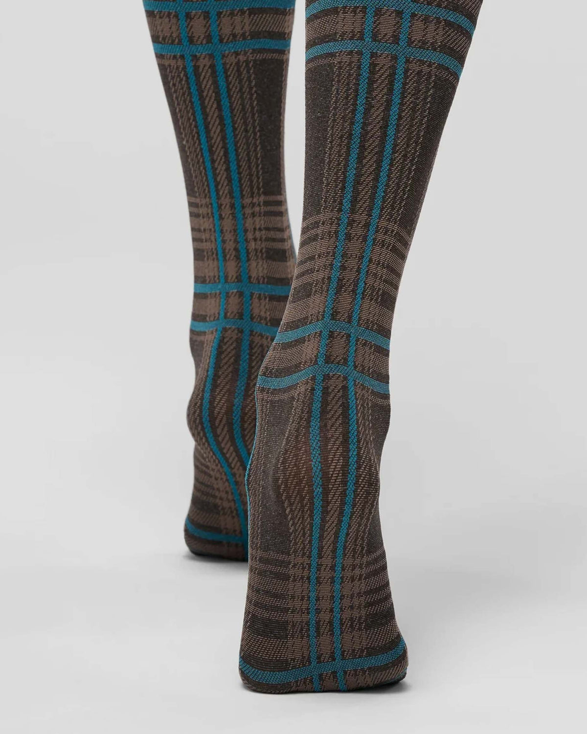 Close up of a pair of knee-high socks with a brown and beige plaid pattern accented by teal blue lines, shown on a model's legs against a light gray studio background.