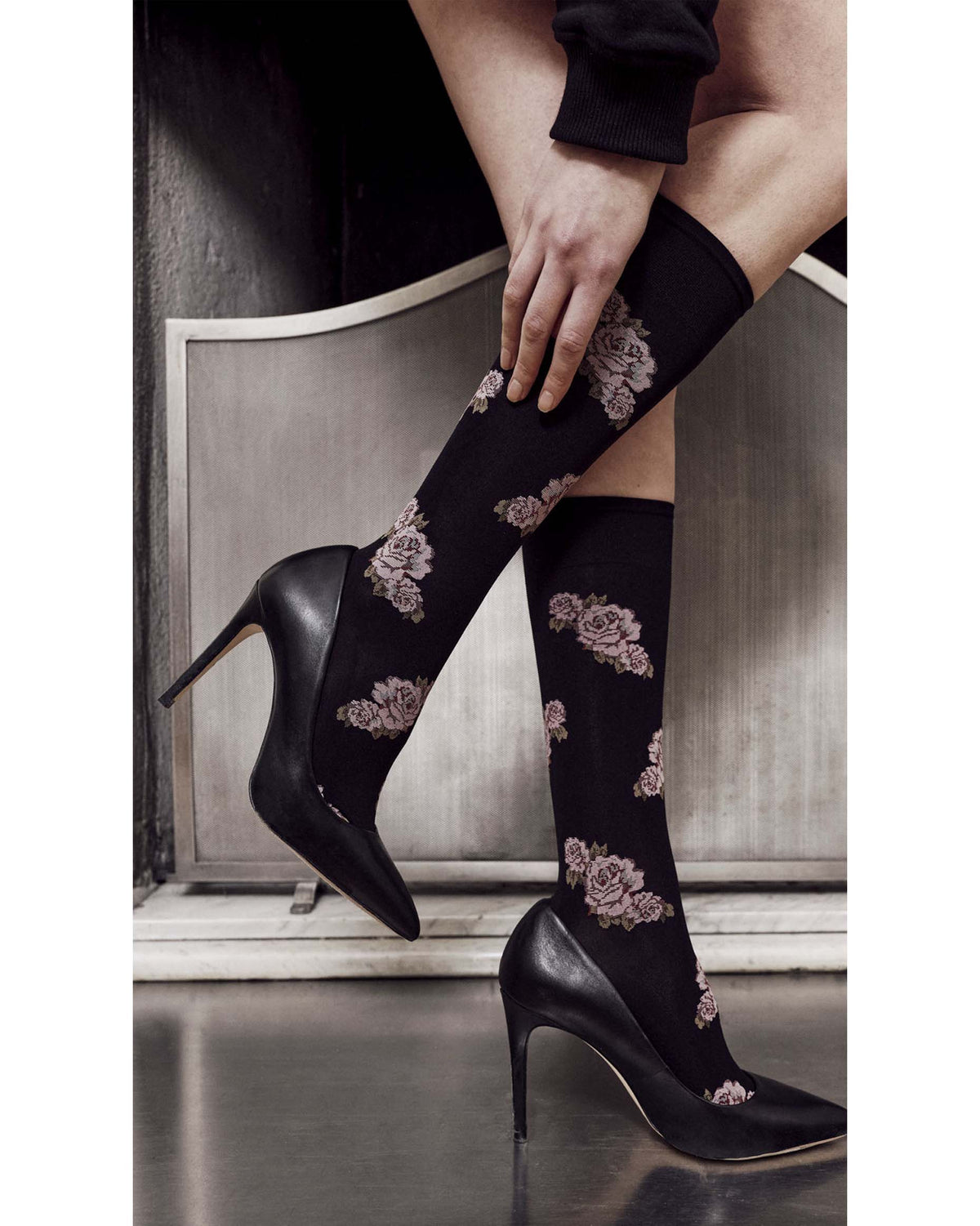A person wearing black knee-high socks with a dusty rose floral pattern, paired with black pointed-toe stiletto heels, posed in front of a gray metal panel.