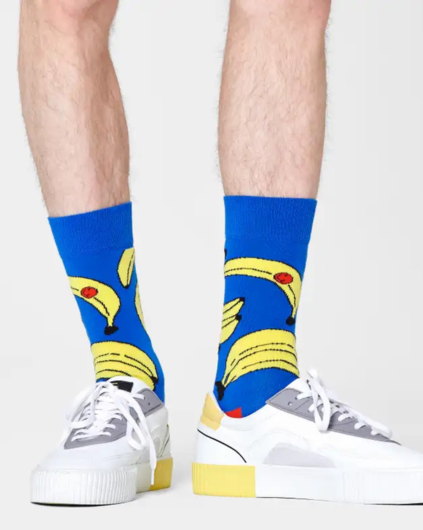 Happy Socks BAN01-6300 Bananas Sock - Bright blue cotton mix crew length ankle socks with a large banana pattern in pale yellow and black with orange smiley face, worn by a man wearing sneakers.