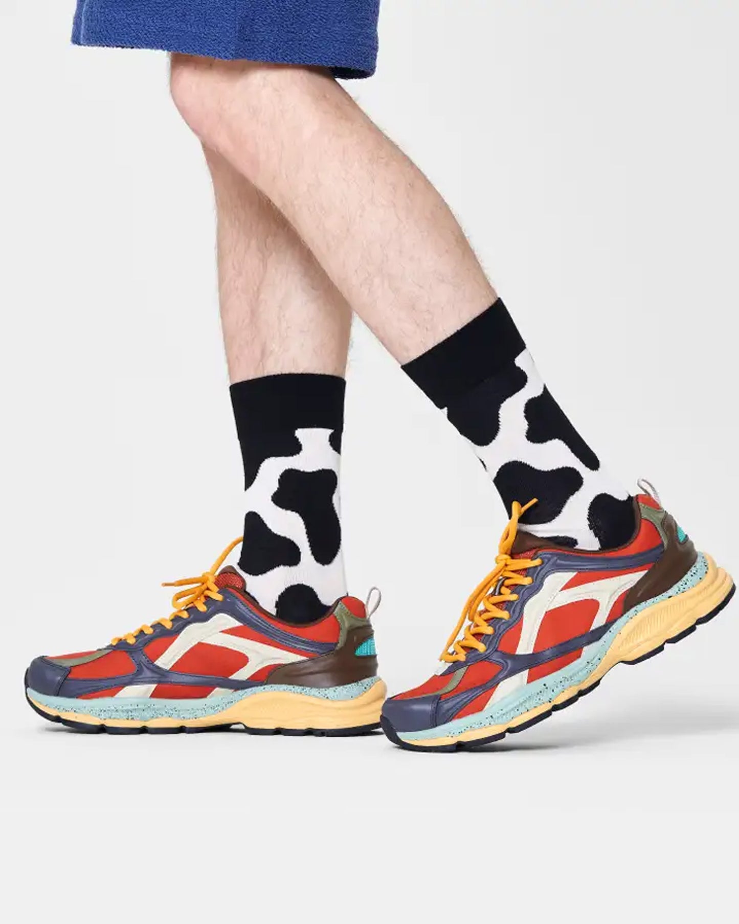 Happy Socks COW01-9300 Cow Sock - Cream cotton mix crew length ankle socks with a black cow print pattern and black cuff, worn by a man wearing trainers and shorts