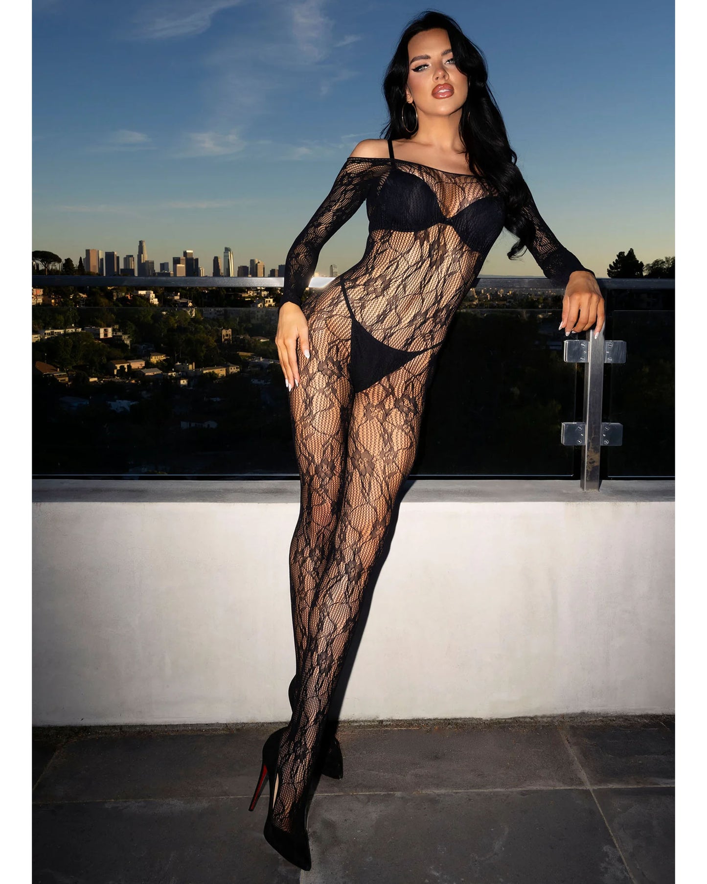 A woman modeling a black long-sleeve fishnet bodysuit with a bra and thong, standing on a rooftop terrace with a city skyline and blue sky in the background.