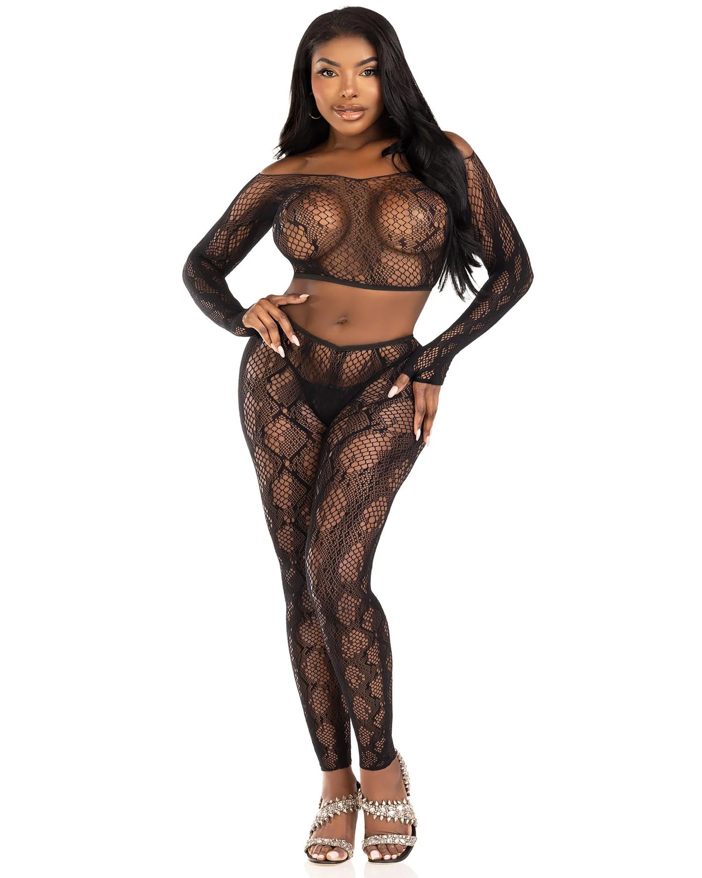 A woman modeling a black fishnet two-piece lingerie set consisting of a long-sleeve crop top, matching high-waisted footless tights, paired with silver studded high-heeled sandals against a plain white background.