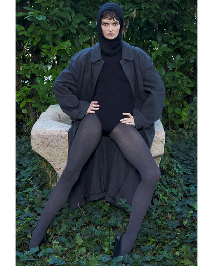 Person wearing a long dark coat, black turtleneck bodysuit and grey ribbed tights sitting on a stone bench with green foliage in the background