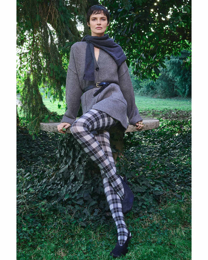 Woman sitting outdoors wearing a gray coat, plaid/tartan tights, and black shoes.