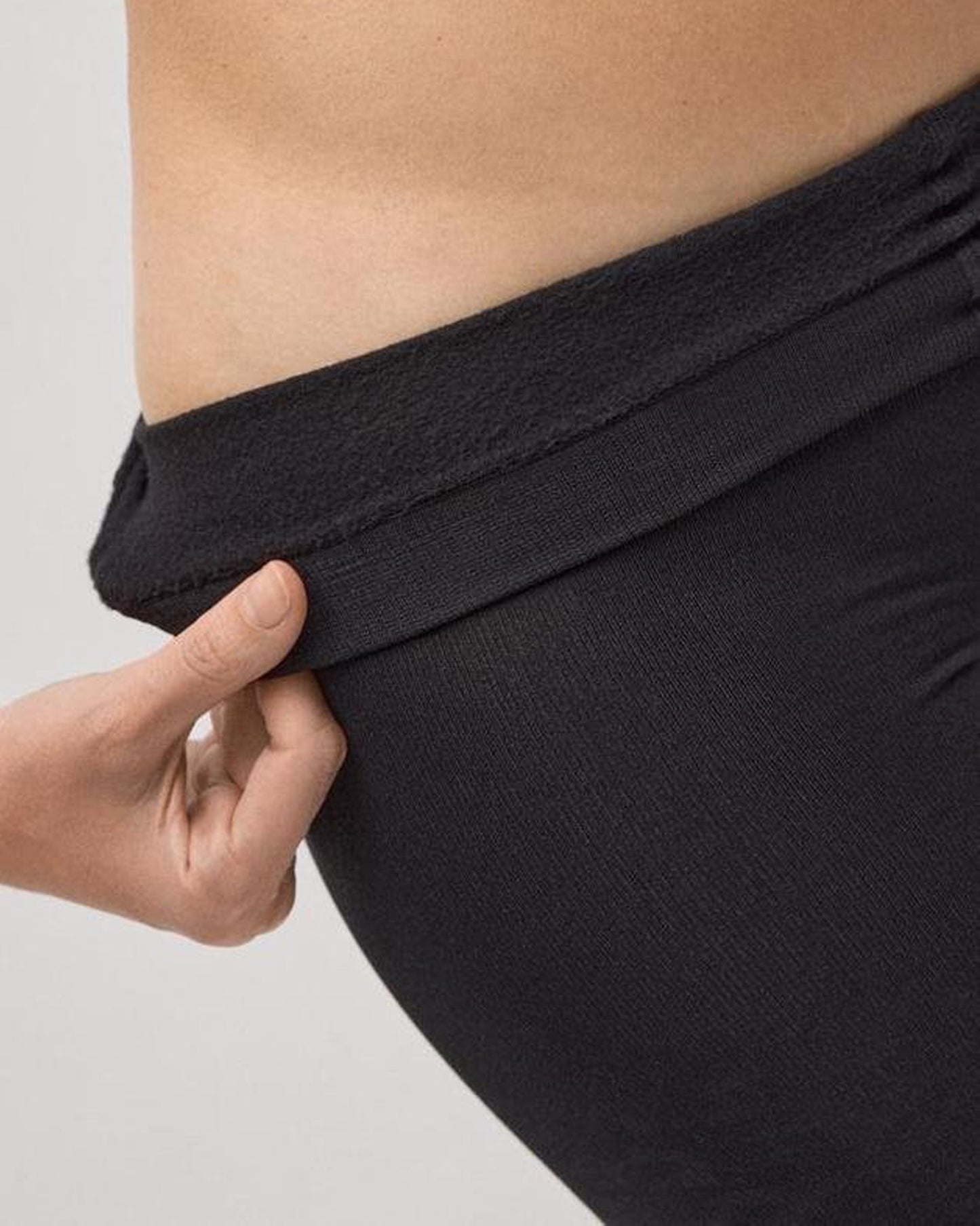 Person folding down the waist band of thermal fleece lined black tights on a plain background