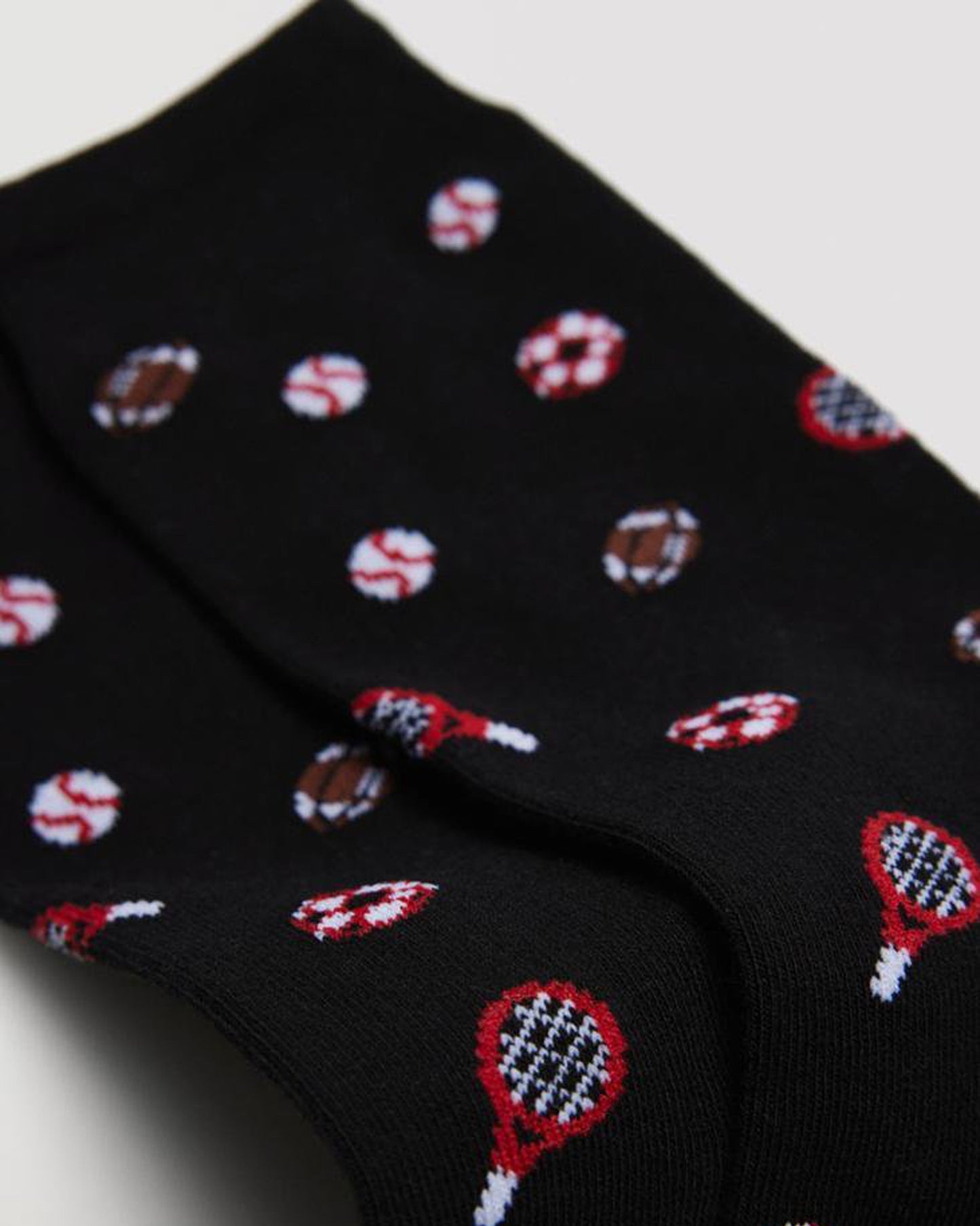 Close up detail of black cotton mix crew length men's ankle socks with an all over pattern of assorted sports balls including soccer, American football, baseball and tennis racket in brown, red and white.