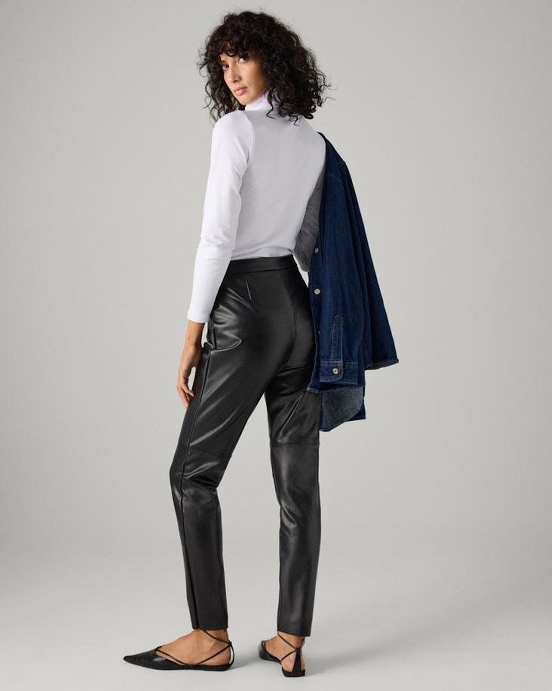 Ysabel Mora 70420 Faux Leather Pants - Black faux leather thermal relaxed fit trouser leggings with fly and button closure, faux front pocket stitching, tapered leg and soft, warm plush fleece lining. Worn with white polo neck. Back view.