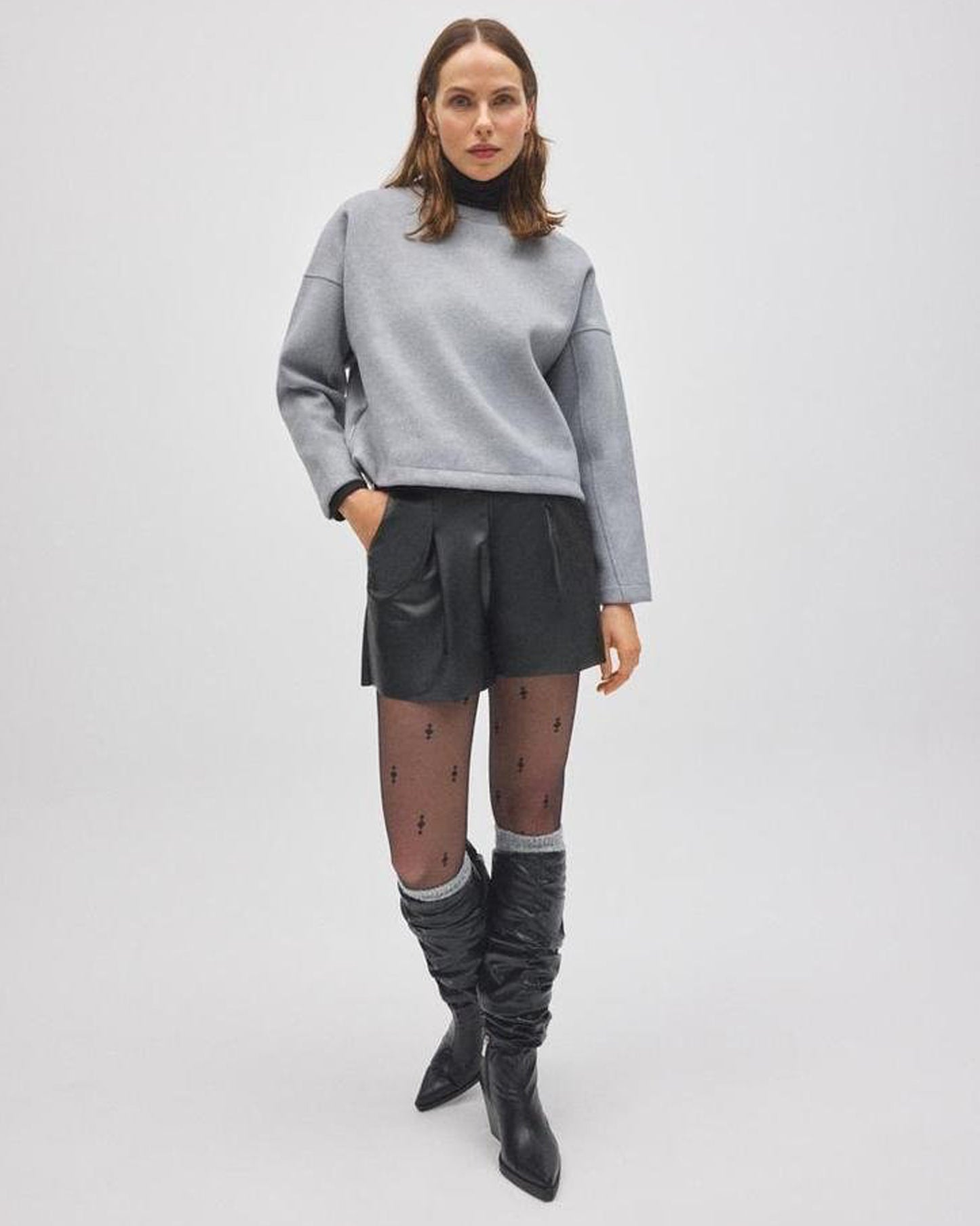Woman wearing a gray sweater, black leather shorts, and gray knee-high socks, black boots on a white background
