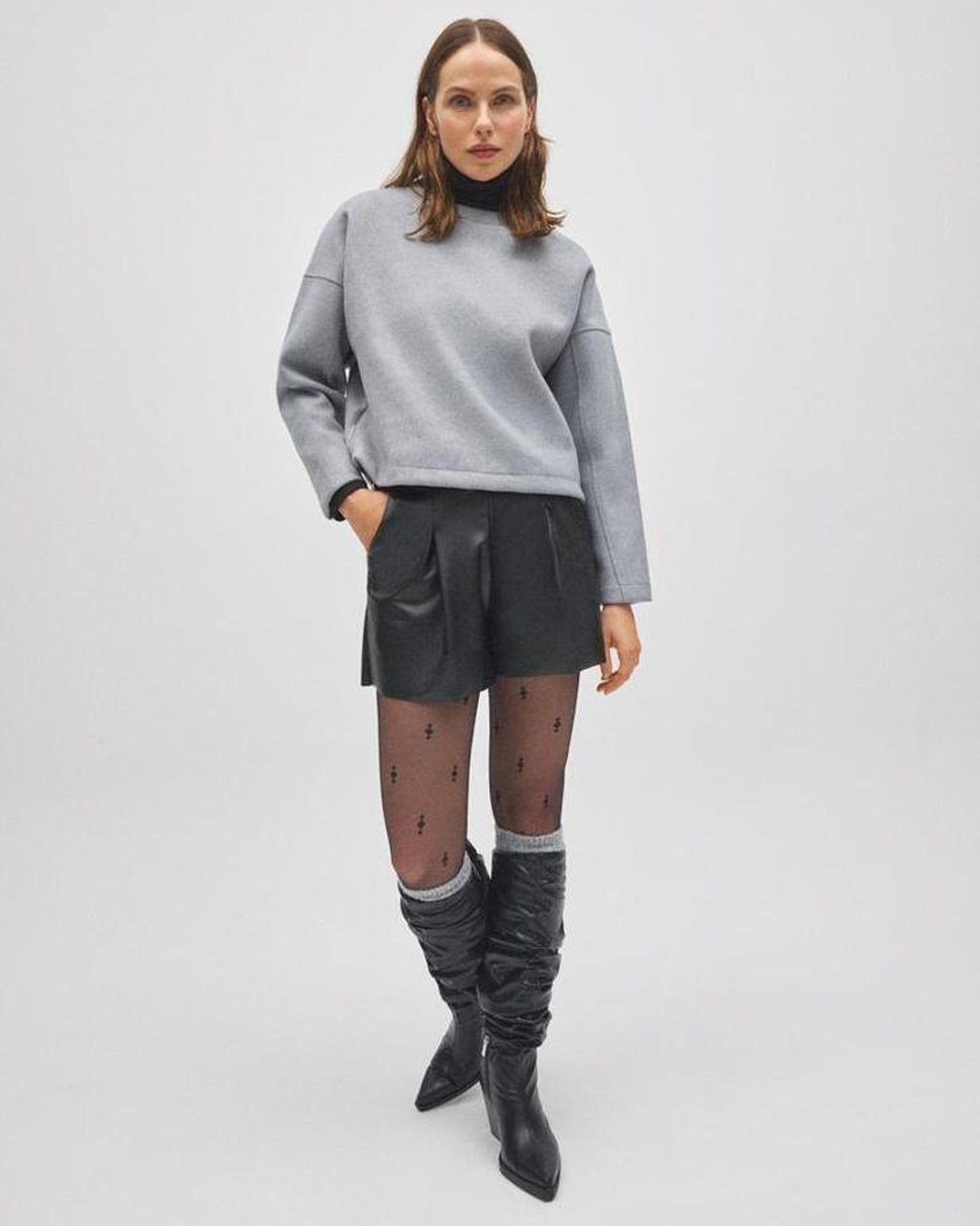 Woman wearing a gray sweater, black leather shorts, and gray knee-high socks, black boots on a white background