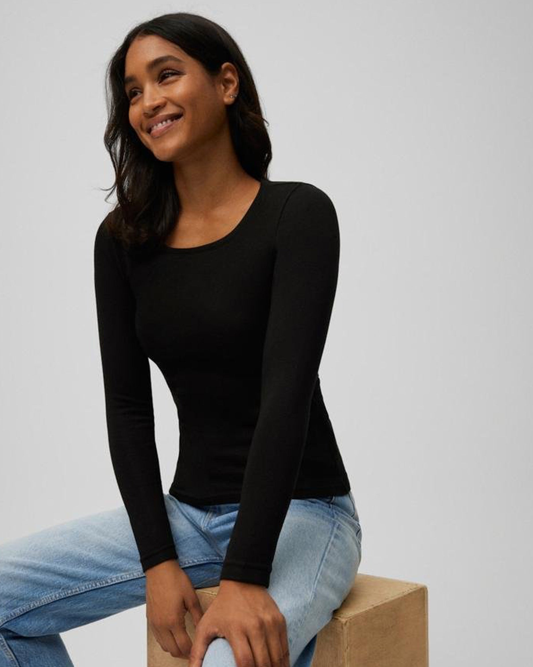 Woman wearing a black long-sleeve top and blue jeans sitting on a beige block against a light gray background
