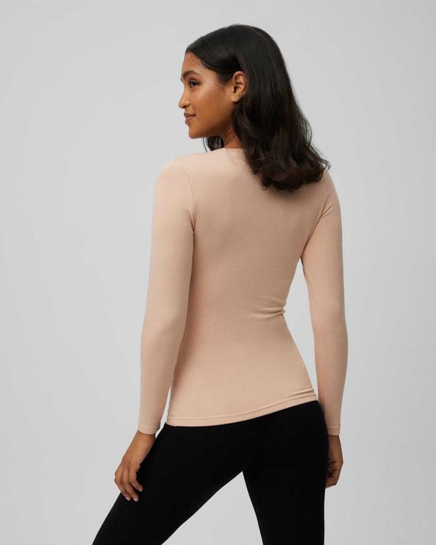 Woman wearing a beige long-sleeve top and black pants on a gray background, back view