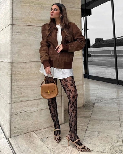 Woman wearing a brown suede jacket , white short dress and black lace patterned tights standing against a building.