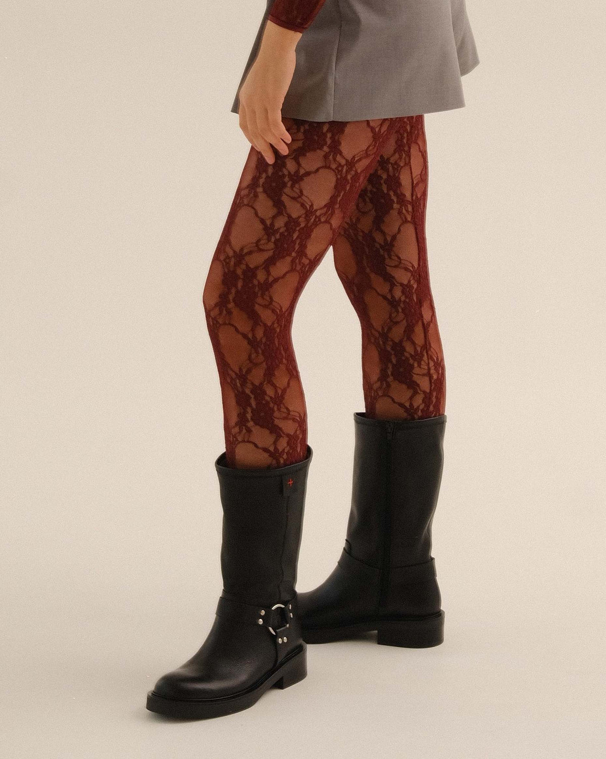 A person wearing sheer burgundy lace tights with a black leather mid-calf boot with a silver buckle, shown from the waist down against a plain light beige studio background.