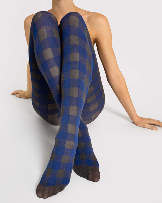 A model seated cross-legged wearing blue and gray buffalo plaid tights against a light gray studio background.