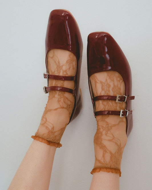 Maroon shoes with buckles worn with sheer beige floral lace socks on a light grey background
