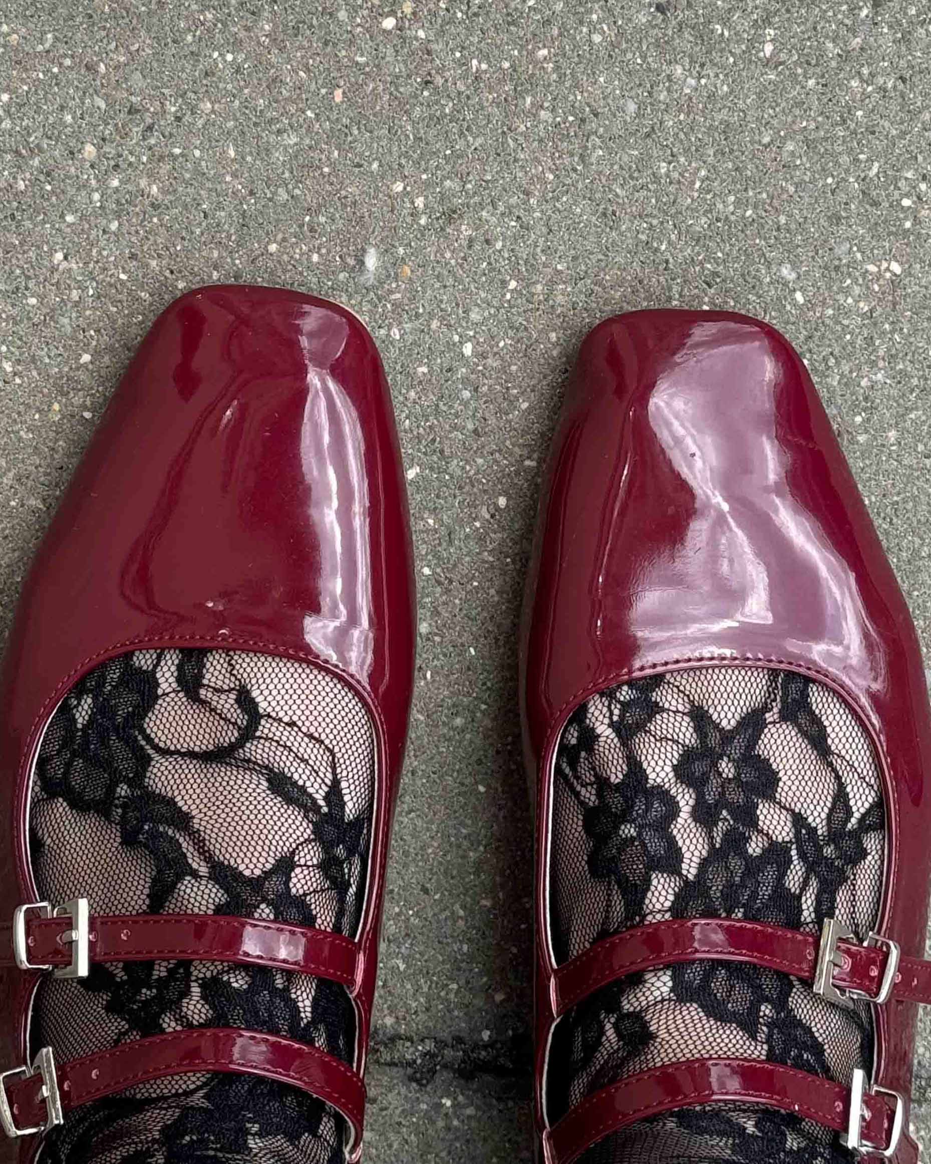 Pair of red patent leather shoes with black lace socks detailing on a grey pavement background