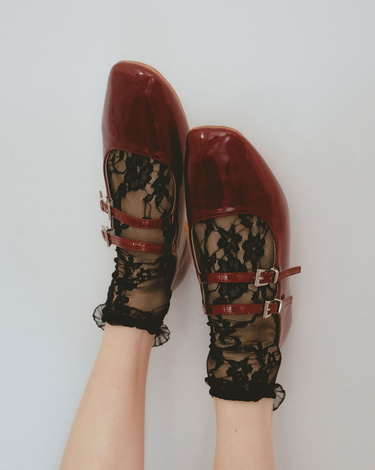 Black floral lace ankle socks worn with red patent leather shoes by a person on a light grey background
