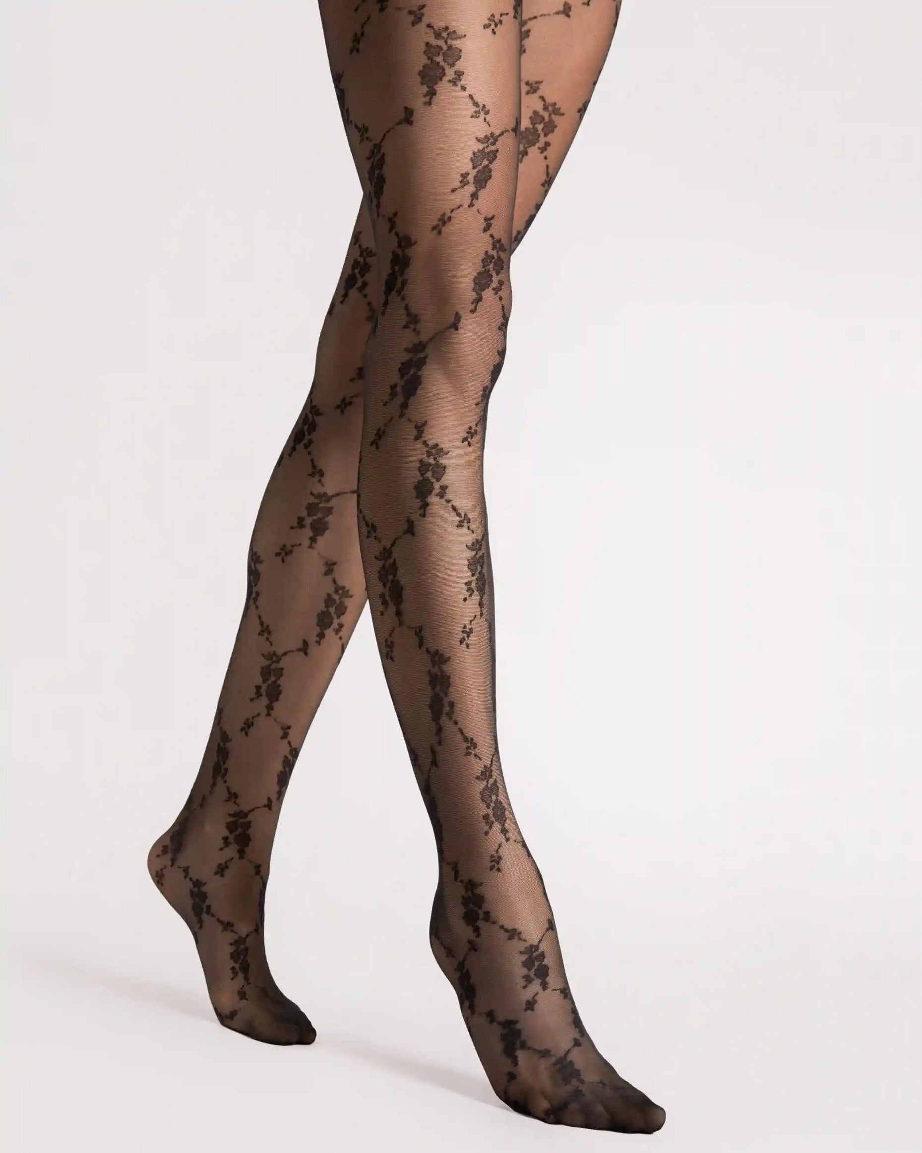 Black floral lace tights worn on legs, shown from the thighs to the feet against a plain light gray studio background.