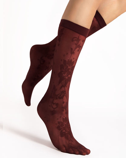 A pair of sheer knee-high socks with a dark floral damask pattern, shown on a model's feet against a clean white background.