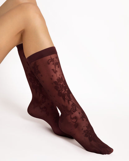 A pair of sheer knee-high socks in a deep burgundy color, featuring an intricate floral lace pattern and a solid ribbed top band, shown on a model's legs against a plain white background.