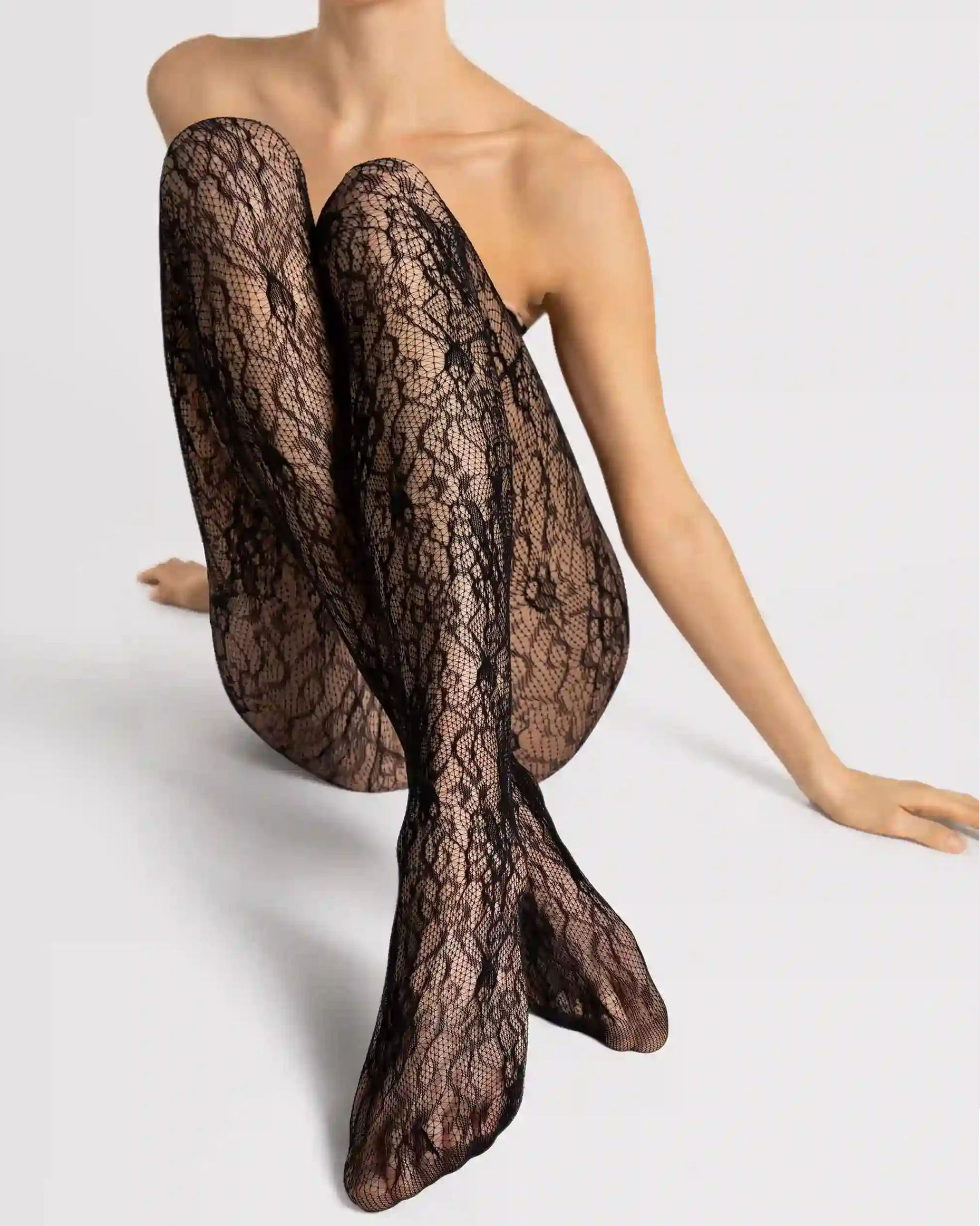 A model wearing sheer black lace tights, posed with legs crossed against a light gray studio background.