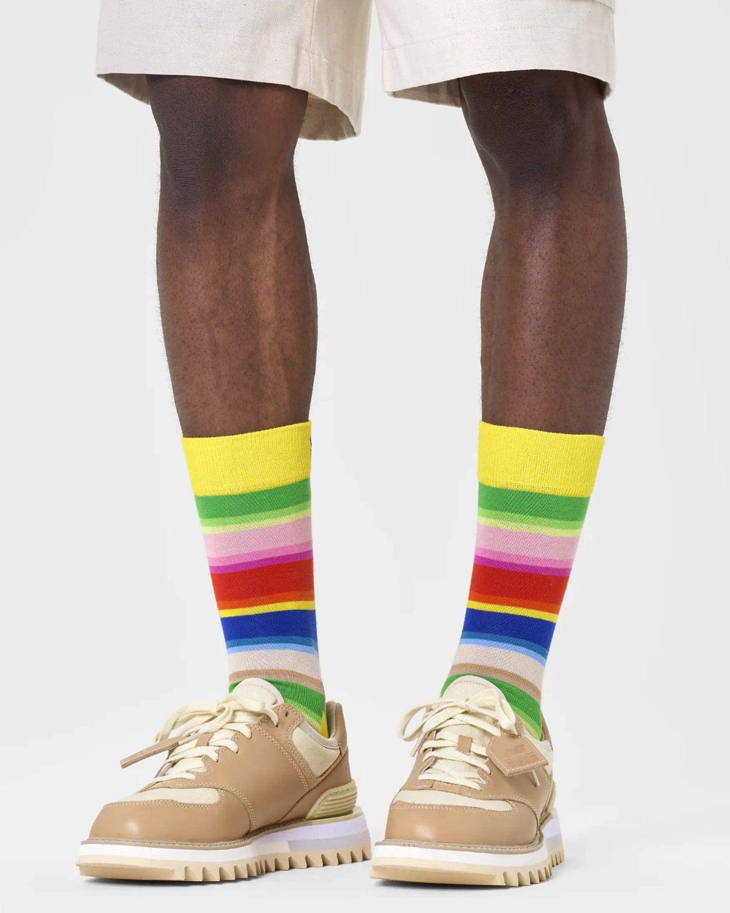 Happy Socks P000834 Gradient Sock - Bright yellow crew length ankle socks with a multicoloured horizontal rainbow stripe pattern. Worn with beige chino shorts and chunky soled tan and cream shoes.