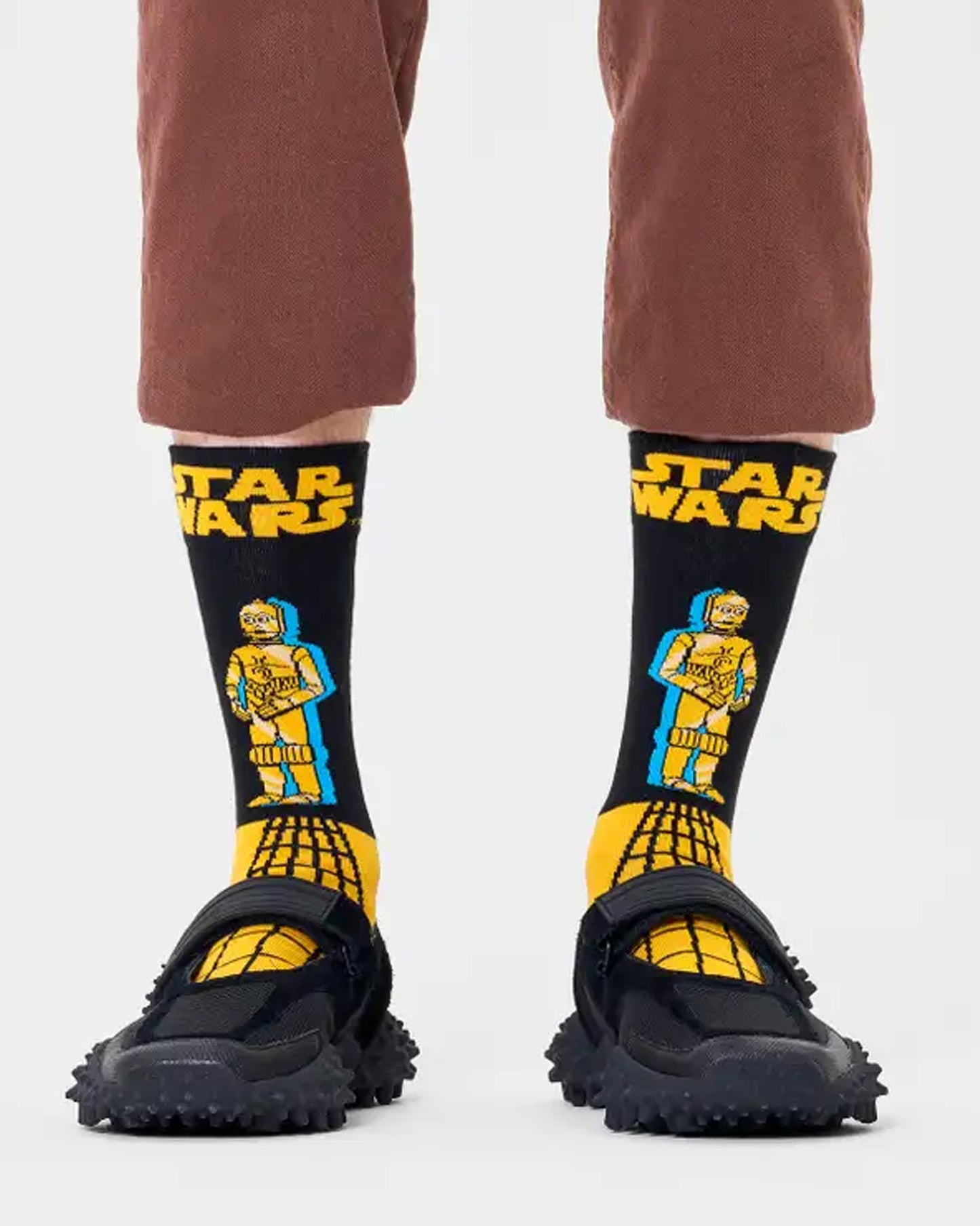 Happy Socks P000273 C-3PO Sock - Star wars themed cotton socks in navy and yellow with C-3PO robot motif.Cotton crew length socks with Star War's logo and C-3PO robot motif in yellow with a turquoise blue shadow on a black background and the bottom half is yellow with a lack linear pattern. Worn with brown cropped pants and slider sandals.