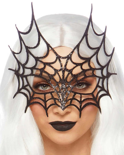 A close-up of a model with long white hair wearing a black spiderweb mask embellished with silver rhinestones, set against a plain white background.