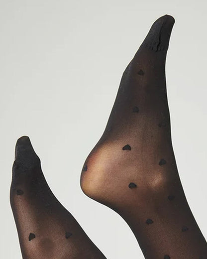 close up of a pair of sheer black tights with small black heart patterns and solid black cuffs, shown on a model's feet against a light gray background.