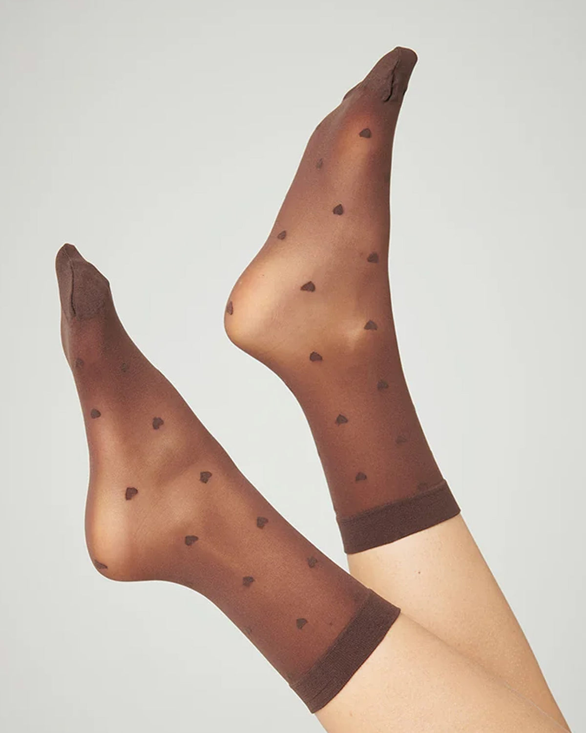 A pair of sheer brown tights with small dark brown heart motifs and solid brown ribbed cuffs, shown on a model's legs against a light gray background.