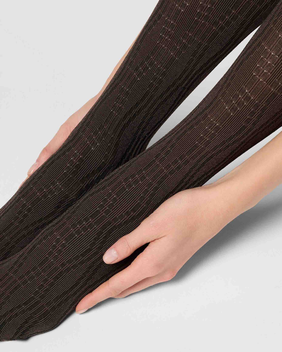 Close-up of a person's legs wearing dark brown ribbed tights with a subtle grid pattern, shown against a light gray background.