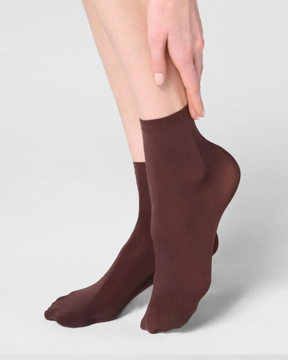 A close-up of a person's lower legs and feet wearing a pair of dark brown ankle socks against a light gray studio background.