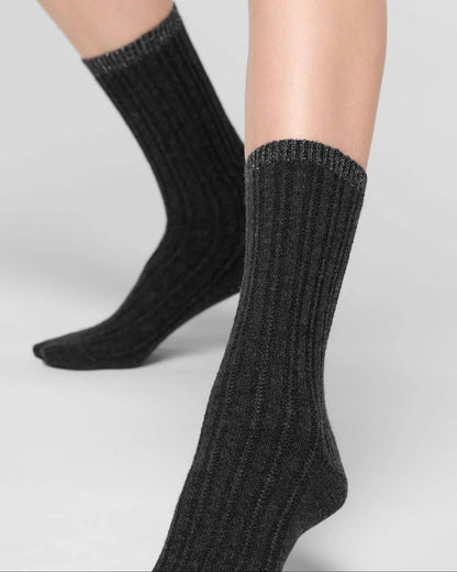 A pair of black ribbed knit crew socks with lamé cuff worn on a model's feet, shown against a neutral light grey studio background.