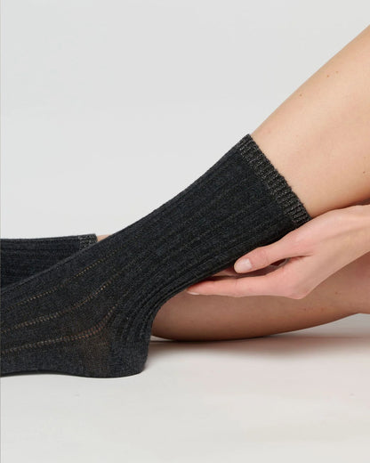 A close-up of a person's foot and lower leg wearing a dark charcoal gray ribbed ankle sock against a light grey studio background.