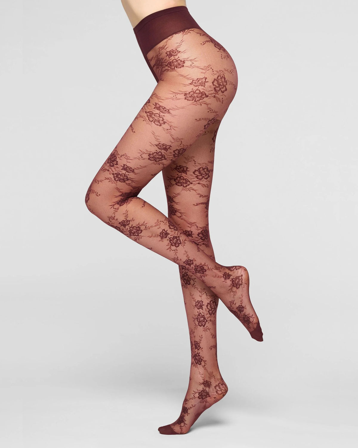 A model's legs wearing sheer rose-beige tights with a dark burgundy floral lace pattern and a matching waistband, shown against a light gray studio background.