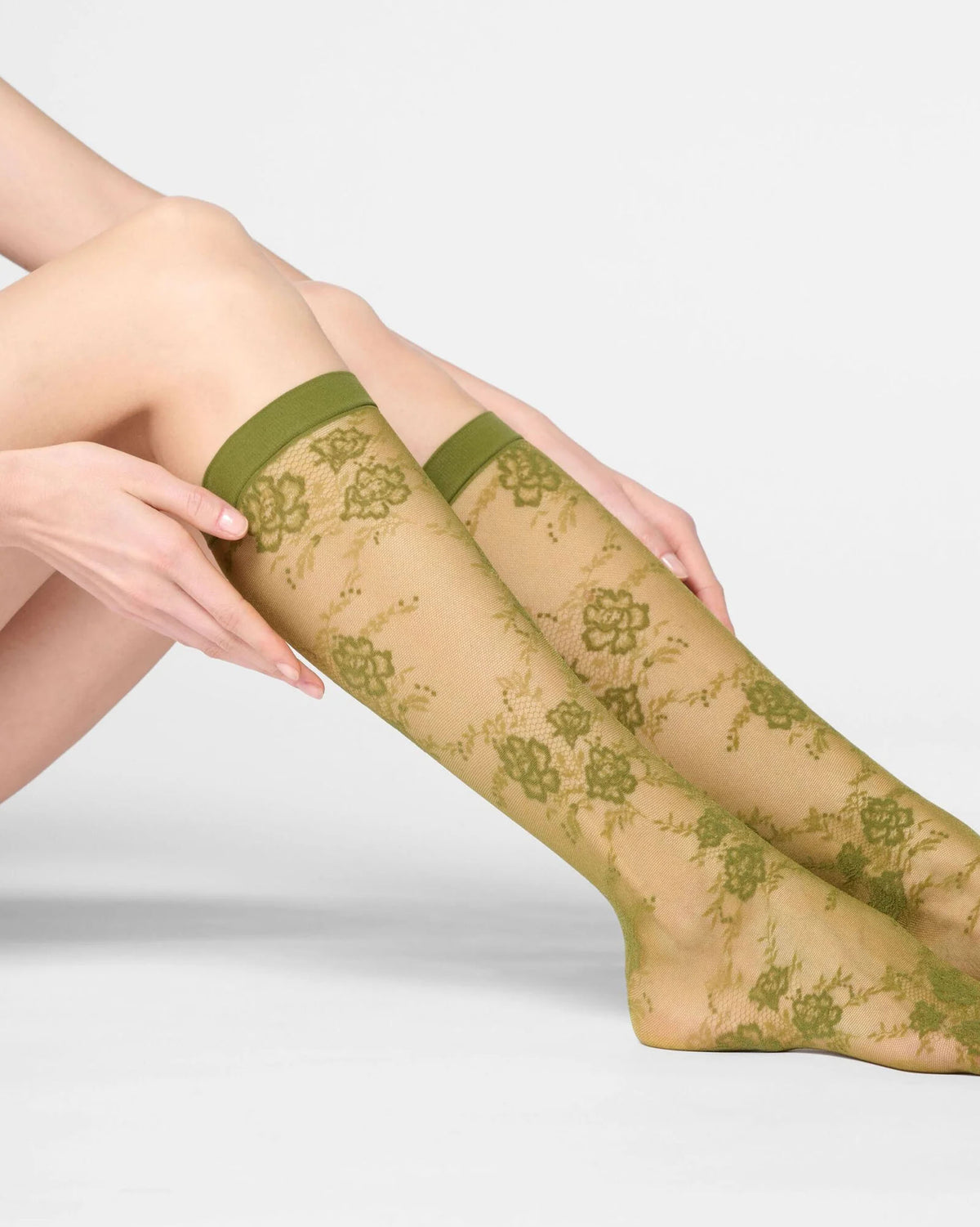 A person seated against a light gray studio background wearing sheer knee-high socks with an olive green floral lace pattern and solid green cuffs.