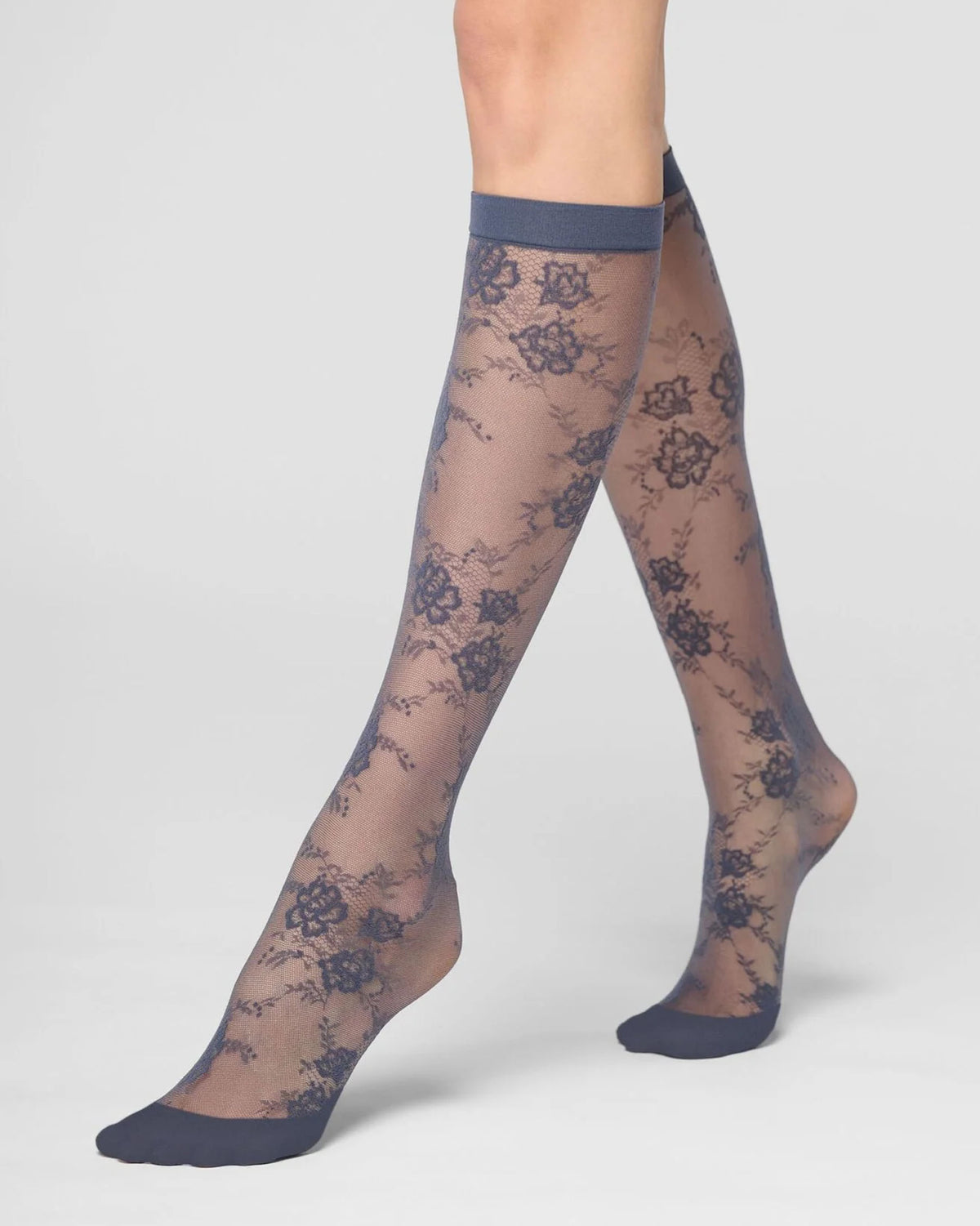 A pair of sheer knee-high socks with a grey blue floral lace pattern and solid cuffs, heels, and toes, shown on a model's legs against a light gray studio background.