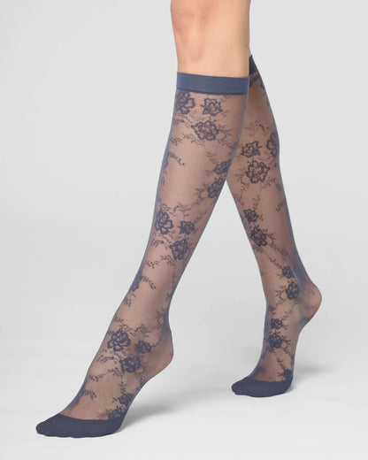 A pair of sheer knee-high socks with a grey blue floral lace pattern and solid cuffs, heels, and toes, shown on a model's legs against a light gray studio background.